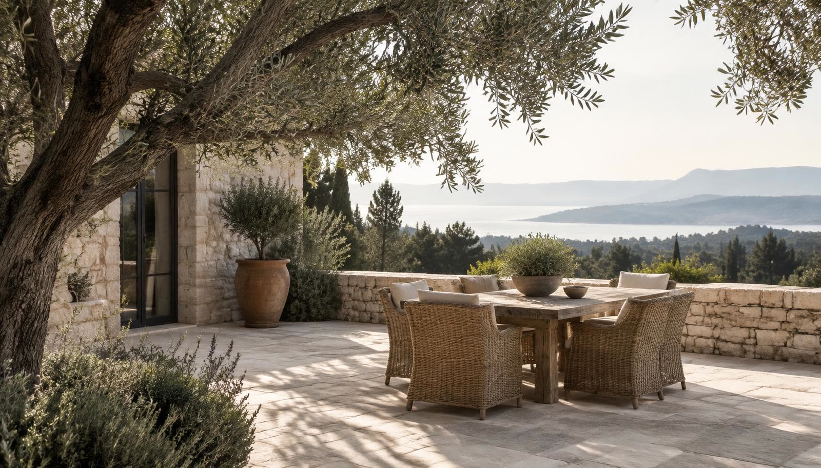 A quiet limestone terrace shaded by an olive tree, with woven chairs around a wooden table and a distant view of the sea in soft morning light.
